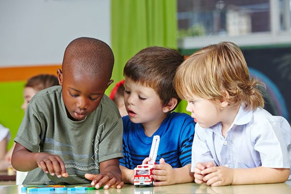 Three children in kindergarten playing with building blocks and cars
